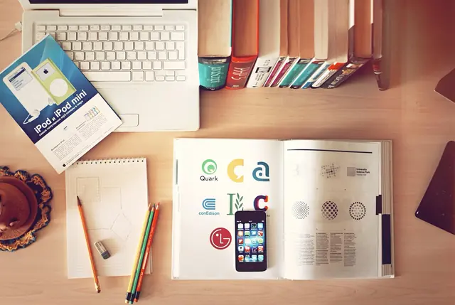 Mesa de trabalho com notebook, lápis, livros e um caderno de referências de logotipos